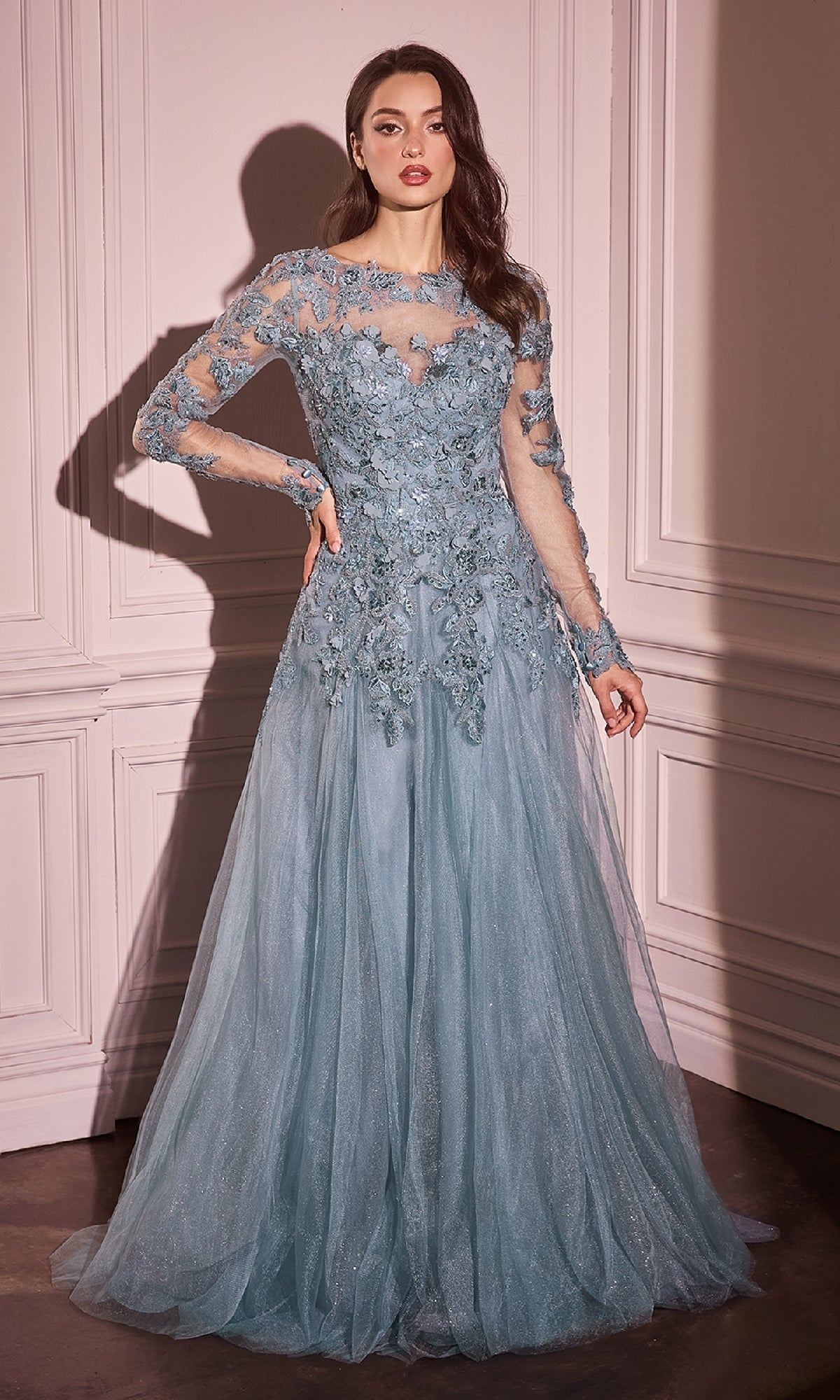 A woman poses indoors against a white paneled wall wearing the Andrea & Leo MA108 long sleeve formal gown in light blue, featuring sheer lace sleeves, floral embroidery, and elegant lace appliques.